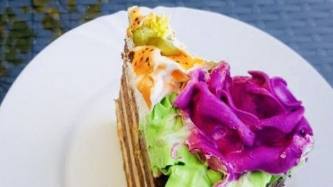 Eine farbenfrohe Stück Buttercreme-Torte liegt auf einem weißen Teller, umgeben von einem transparenten, reflektierenden Untergrund.