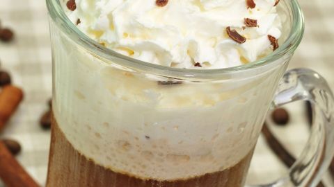 Ein Glas mit schaumiger Moose Milk, dekoriert mit Sahne und Schokoladenstückchen, steht auf einer karierten Tischdecke umgeben von Zimtstangen und Kaffeebohnen.