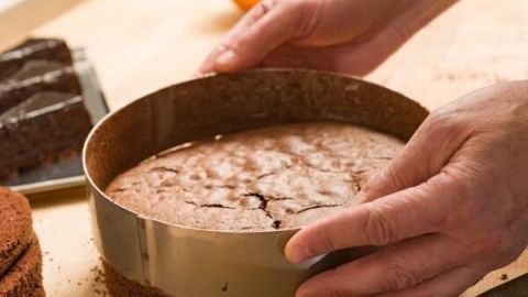 Hände halten einen frisch gebackenen Kuchen ohne Ei in einer Form auf einem Holztisch, umgeben von Küchenutensilien.
