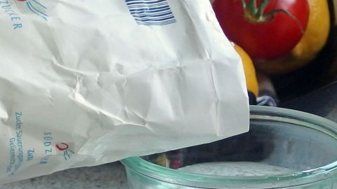 Zucker wird in ein Glas mit Wasser gegossen, um Fruchtfliegen anzulocken, während frisches Obst im Hintergrund liegt.