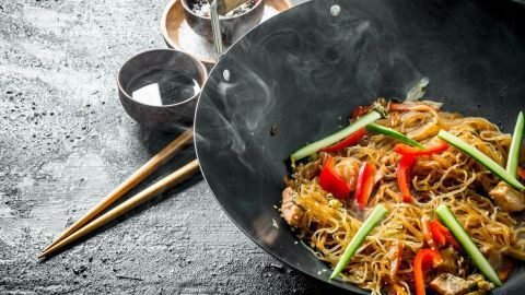Bunte Asia-Pfanne mit gebratenen Nudeln und Gemüse, dampfend in einem Wok, auf einer dunklen Oberfläche.