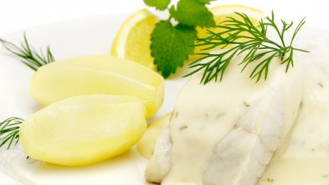 Auf dem Teller liegt ein Karpfenfilet in cremiger Soße, umgeben von Kartoffeln und frischen Kräutern, Zitrone ist dekorativ platziert.