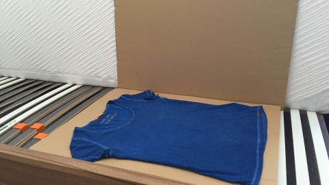 Ein blauer T-Shirt liegt auf einer Pappunterlage auf einem Bett mit gestreiftem Bezug, bereit zum Bügeln.