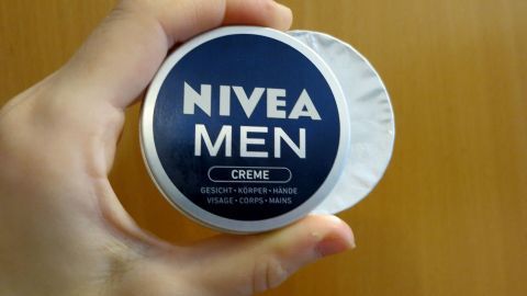 Eine Hand hält eine runde Dose mit Nivea Men Creme, die zur Hautpflege verwendet wird, vor einer Holzoberfläche.