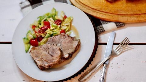 Ein Salzbraten auf grünem Teller mit Kartoffelsalat und Tomatenscheiben, daneben ein Brotmesser auf Holztisch.