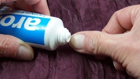 Eine Hand drückt Zahnpasta auf die Finger, die weiße Spitzen haben, auf einer texturierten lila Unterlage.