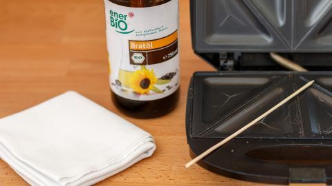 Ein Sandwichtoaster steht geöffnet auf einem Holztisch, daneben sind ein Putzlappen, ein Holzstab und eine Flasche Reinigungsmittel.