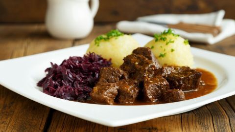Saftiges Hirschgulasch in einer geschmackvollen Soße, begleitet von Rotkohl und Kartoffelknödeln auf einem weißen Teller.