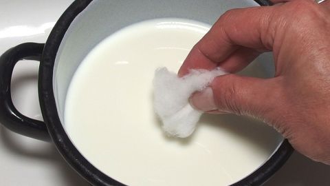 Gegen Augenringe hilft es einen mit Milch getränkten Wattepad auf die Augen zu legen. Eine Hand taucht ein Wattepad in eine Schüssel mit Milch, um es gegen Augenringen zu verwenden.