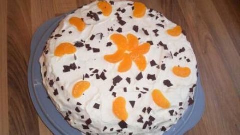 Eine dekorierte Zitronen-Mandarinen Torte liegt auf einem blauen Teller, umgeben von einem Holztisch. Schokoladenstückchen und Mandarinen zieren die Sahne.
