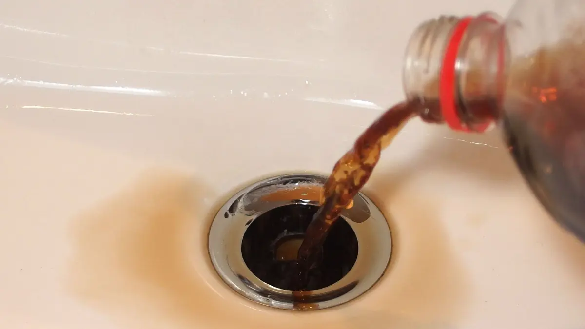 Eine Flasche Cola wird in ein Abflussloch gegossen, w&auml;hrend die braune Fl&uuml;ssigkeit durch den Abfluss flie&szlig;t.
