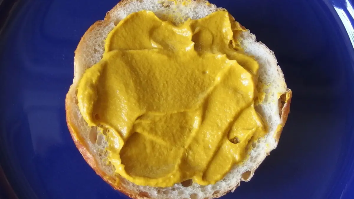Ein Br&ouml;tchen mit gleichm&auml;&szlig;ig aufgetragenem Senf liegt auf einem blauen Teller, bereit zum Verzehr.