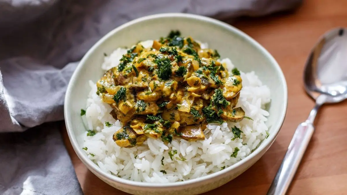Ein Teller mit Reis, belegt mit einer w&uuml;rzigen Champignon-Curry-Sahne-Mischung, steht auf einem Tisch mit einer silbernen Gabel daneben.