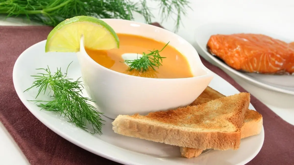 Eine cremige Lachscremesuppe in einer wei&szlig;en Schale, garniert mit Dill und Zitrone, serviert mit Toast auf dem Teller.