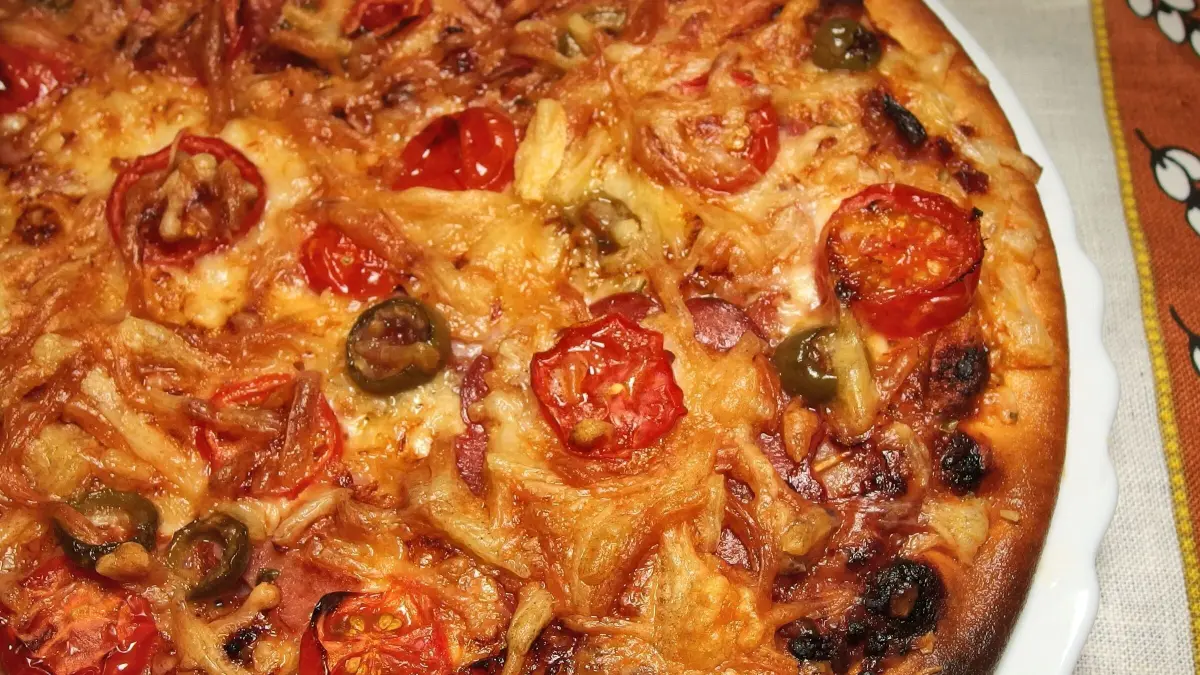Eine frisch gebackene Pizza mit Tomaten und gr&uuml;nen Oliven liegt auf einem Teller, bereit zum gemeinsamen Essen beim Kindergeburtstag.