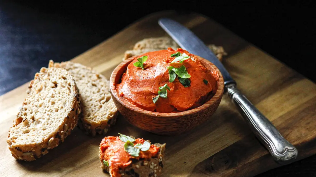 Eine Sch&uuml;ssel mit Tomaten-Basilikum-Butter steht auf einem Holzbrett neben geschnittenem Vollkornbrot.