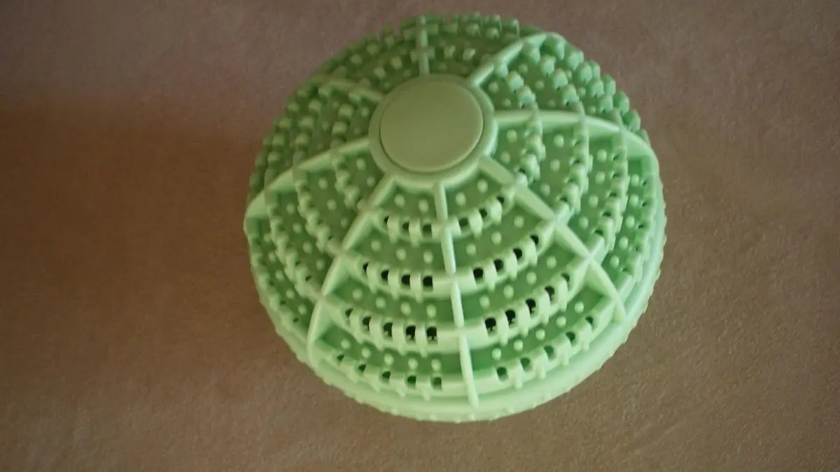 Ein gr&uuml;ner &Ouml;ko-Waschball liegt still auf einer beige Oberfl&auml;che. Sein Design ist strukturiert und rund.