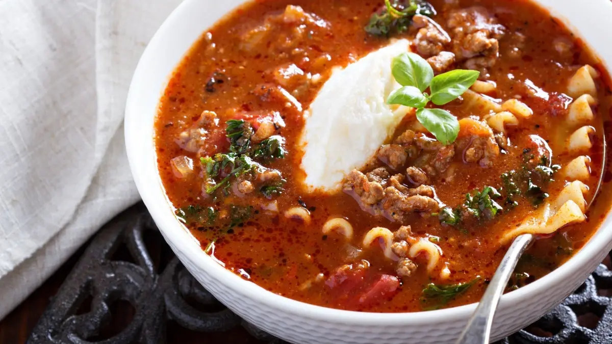 Eine dampfende Partysuppe mit Hackfleisch und Kr&auml;utern in einer Sch&uuml;ssel, garniert mit einem L&ouml;ffel Sahne und Basilikum.