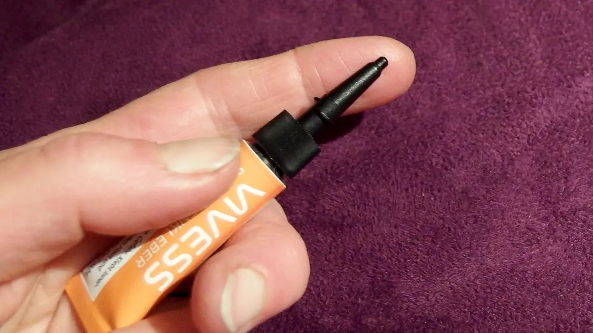 Eine Hand h&auml;lt eine Tube Sekundenkleber, w&auml;hrend der Hintergrund lila ist und die Finger vorsichtig positioniert sind.