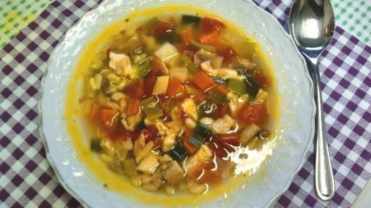 Eine Sch&uuml;ssel Nudelsuppe mit H&uuml;hnerfleisch, Gem&uuml;se und Br&uuml;he, auf einem karierten Tischl&auml;ufer serviert, neben einem L&ouml;ffel.