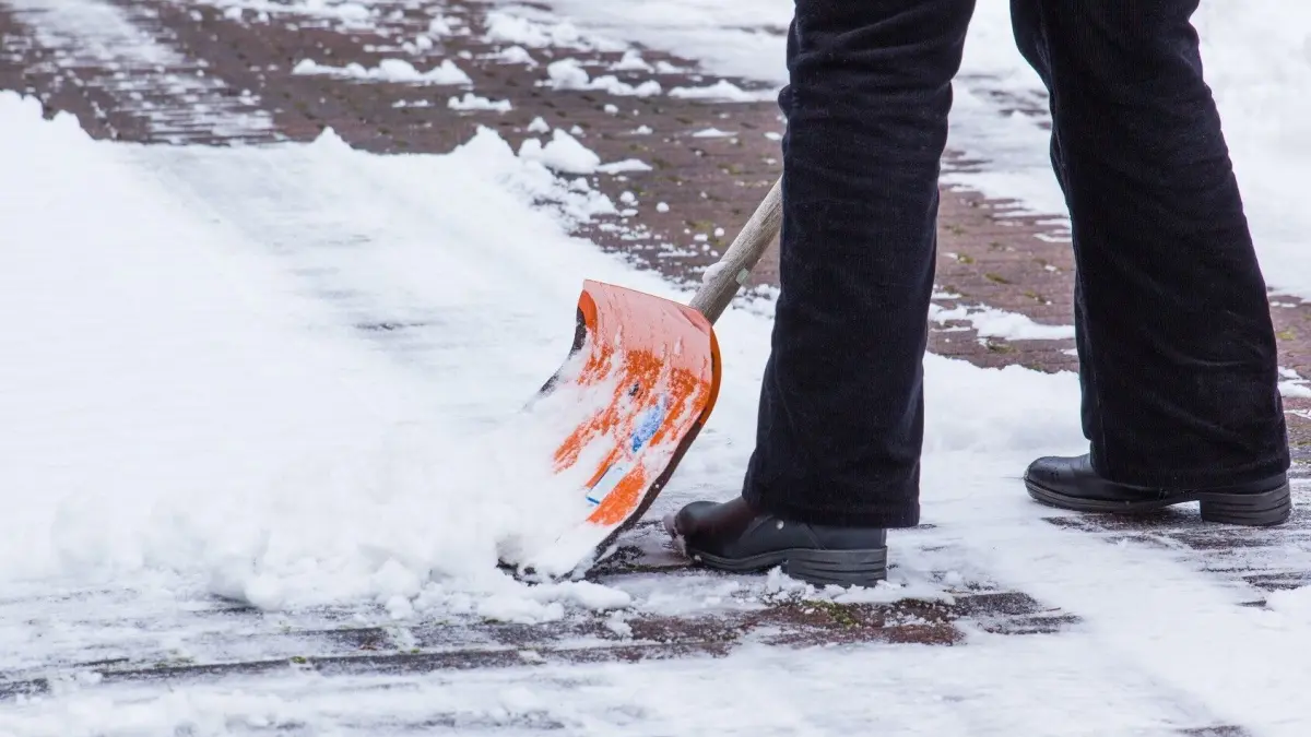 Jemand schaufelt Schnee auf einem Gehweg, tr&auml;gt rutschfeste Schuhe und arbeitet bei Schneematsch und Gl&auml;tte.