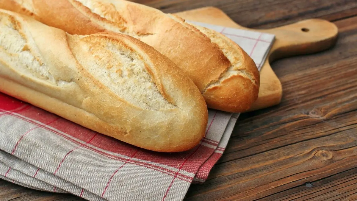 Zwei frisch gebackene Wei&szlig;brote liegen auf einem Holzbrett, umgeben von einem karierten Geschirrtuch auf einem Holztisch.