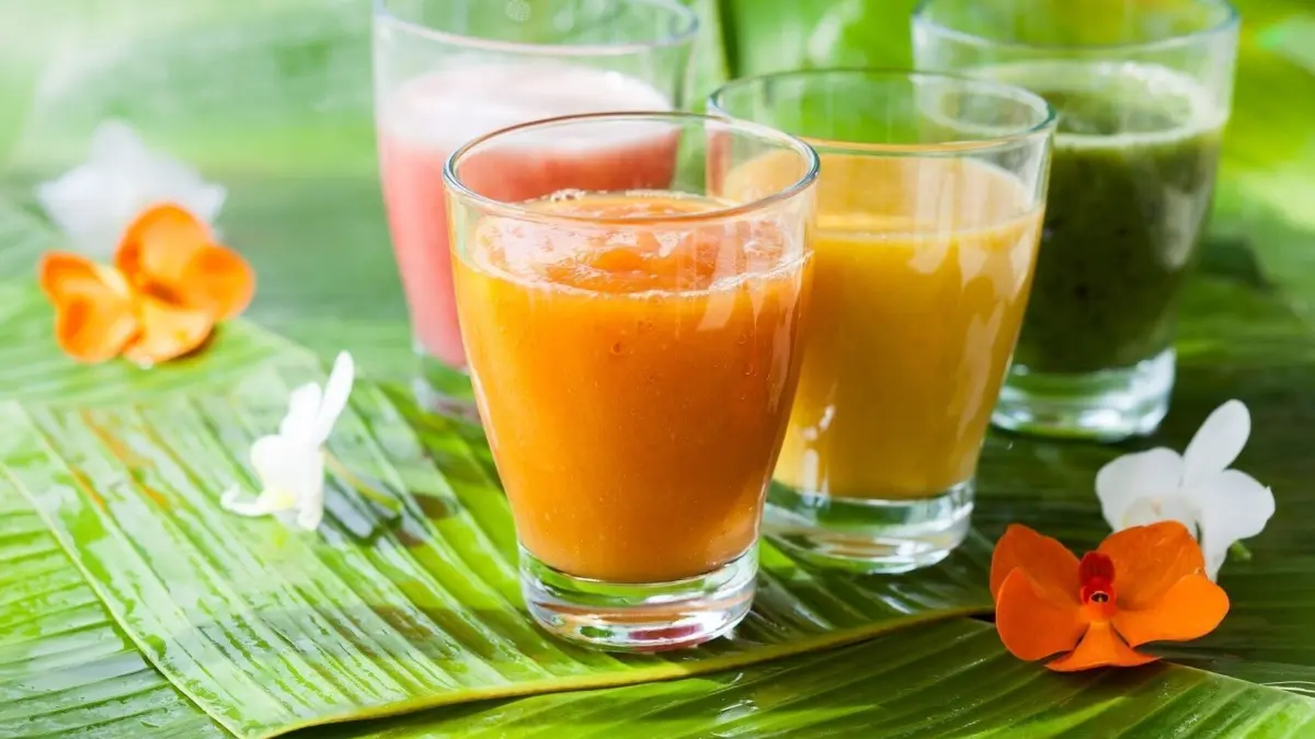 Bunte Smoothies stehen in Gl&auml;sern auf gr&uuml;nen Bl&auml;ttern, umgeben von dekorativen Blumen, die frische gesunde Genussmomente ausstrahlen.