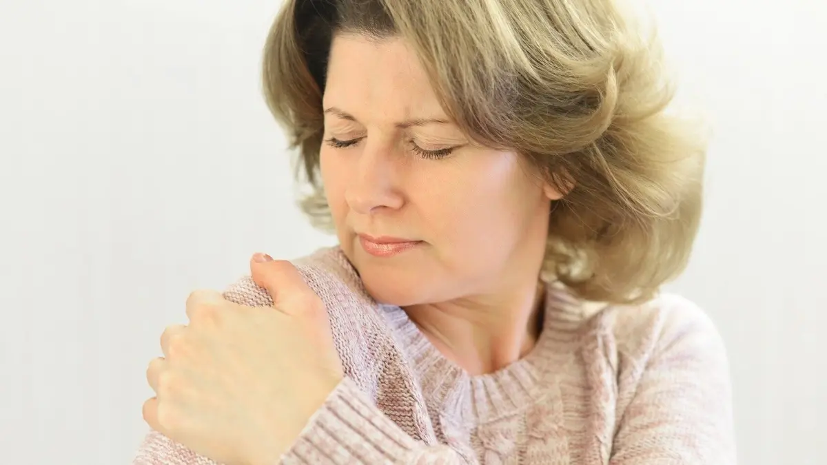 Eine Frau greift schmerzerf&uuml;llt an ihre Schulter, was auf eine Frozen Shoulder hindeutet, in einem hellen Raum.