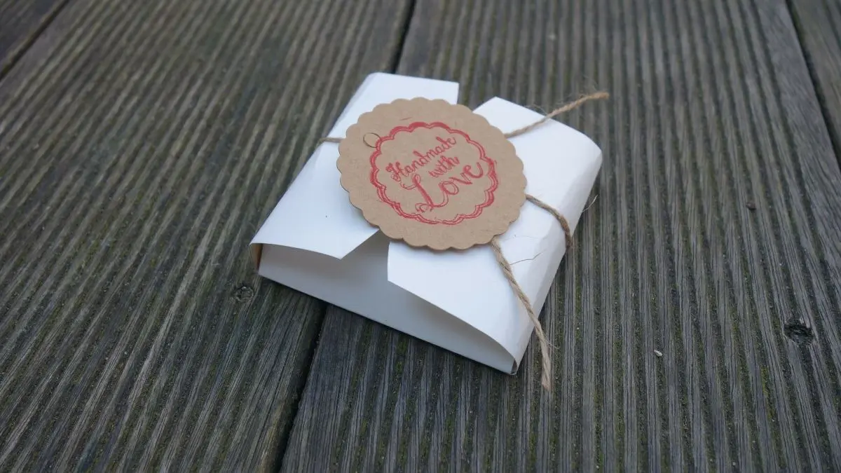 Eine dekorative Keksschachtel aus Papier liegt auf einer Holzoberfl&auml;che, elegant mit einer Schnur und einem Anh&auml;nger verziert.