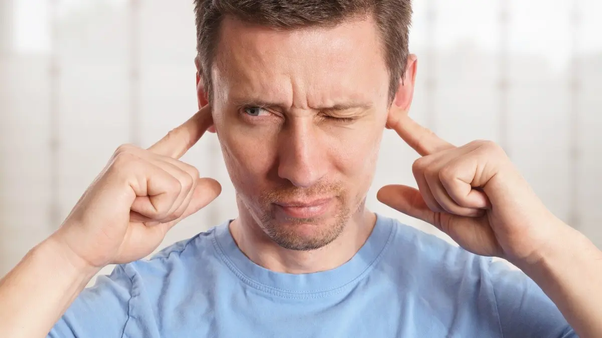 Ein Mann h&auml;lt sich die Ohren zu, w&auml;hrend er eine schmerzhafte Miene macht, als w&uuml;rde er unter Tinnitus leiden.
