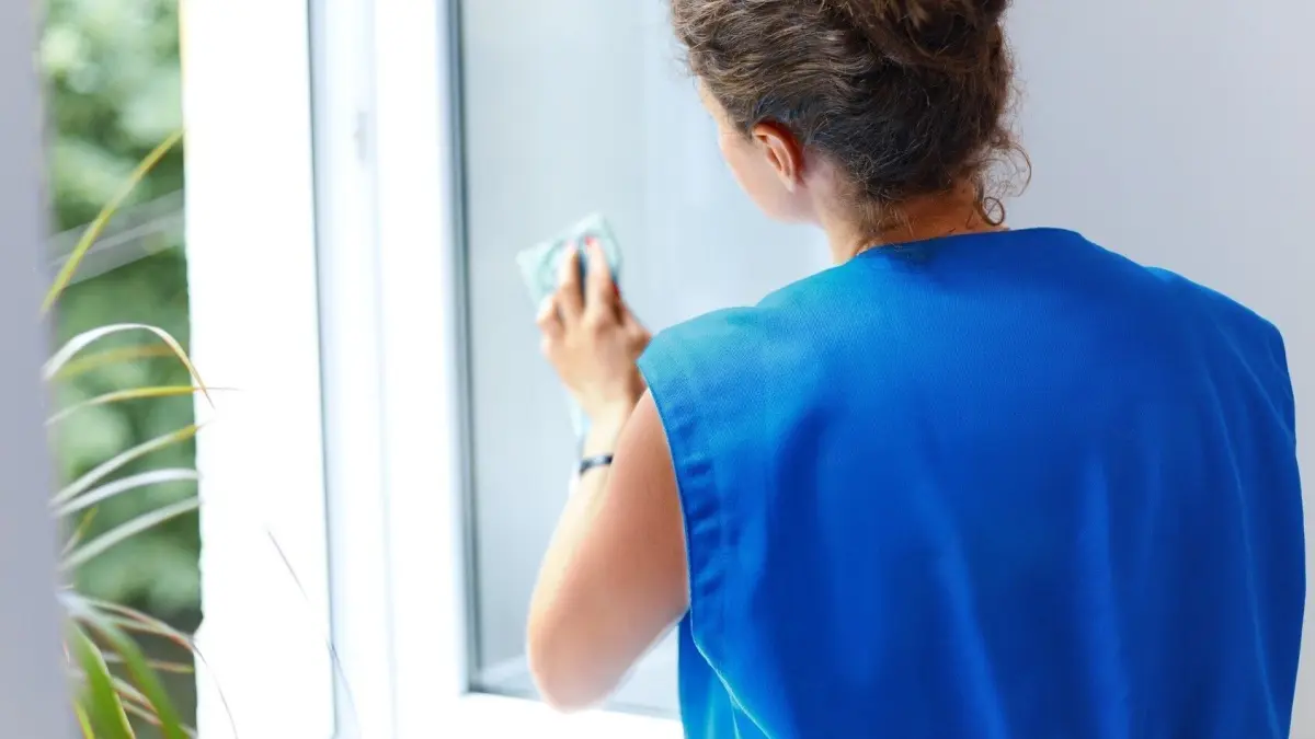 Eine Person in blauer Kleidung putzt streifenfrei ein Fenster mit einem Reinigungstuch in einem hellen Raum.