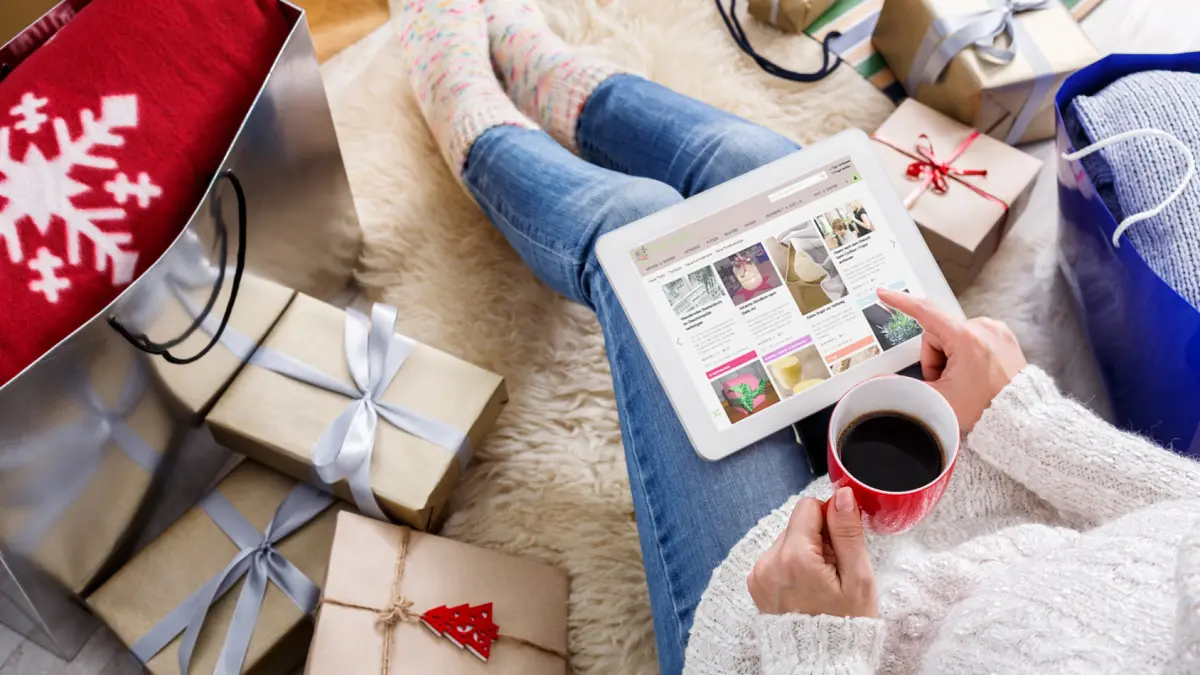Eine Person sitzt auf einem Teppich, blickt auf ein Tablet und genie&szlig;t einen warmen Tee, umgeben von Weihnachtsgeschenken.