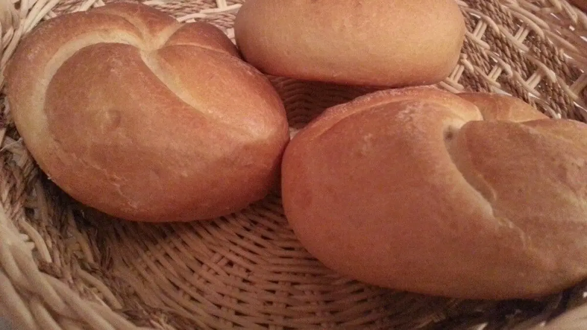 Drei goldbraune Br&ouml;tchen liegen frisch gebacken in einem geflochtenen Korb auf einem Tisch.
