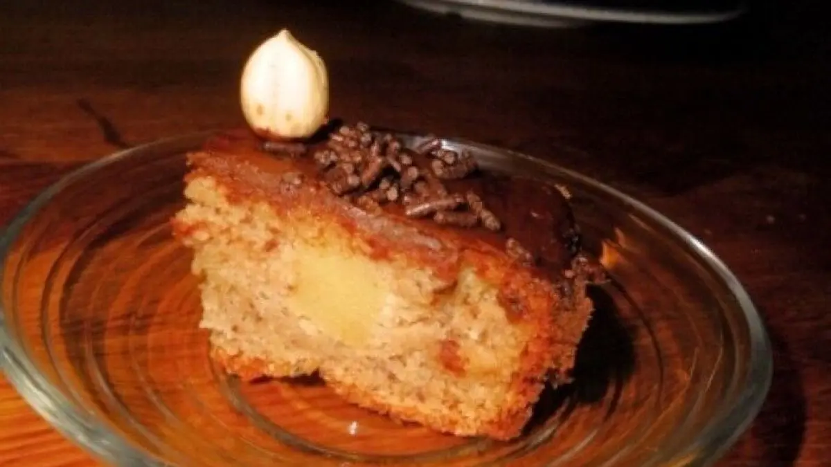 Ein St&uuml;ck Nusstorte liegt auf einem Glasuntersetzer, w&auml;hrend im Hintergrund eine dreist&ouml;ckige Torte sichtbar ist.