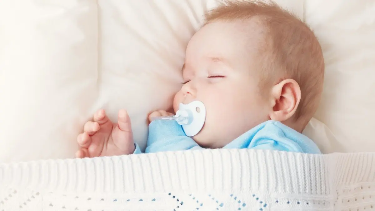 Ein schlafendes Baby mit einem Schnuller liegt geborgen in einer Decke auf einem weichen Kissen, in ruhiger Umgebung.