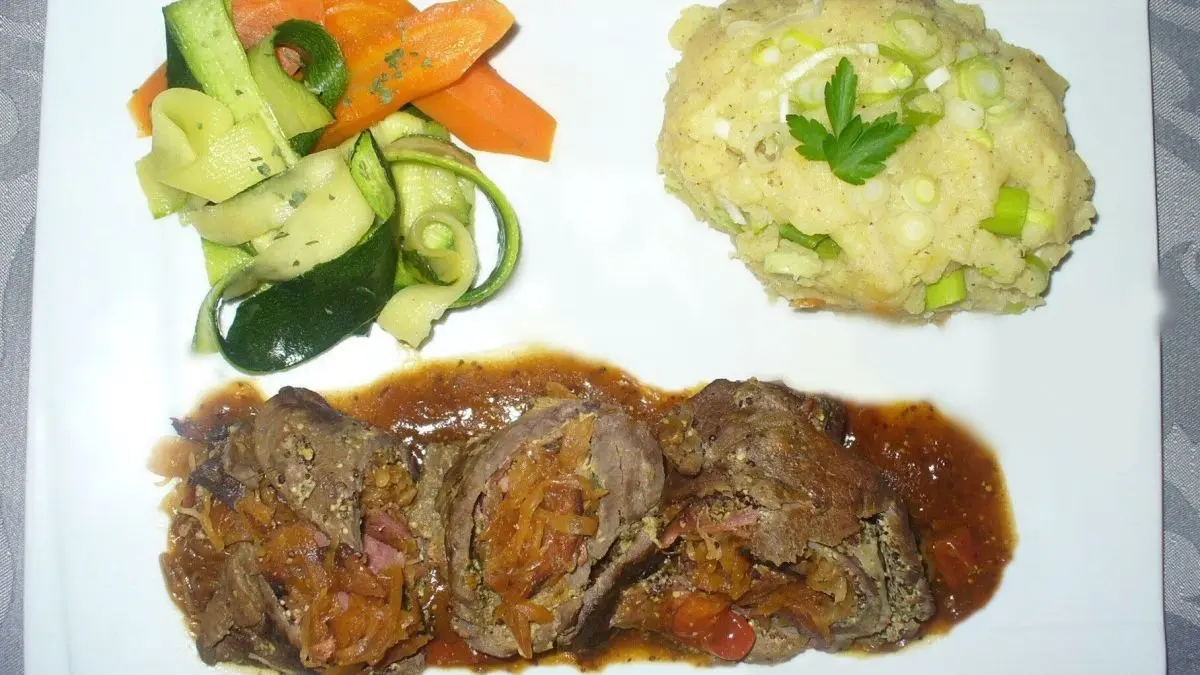 Zarte Rinderrouladen mit Sauce liegen auf einem wei&szlig;en Teller, umgeben von Gem&uuml;se und Kartoffelbrei, appetitlich angerichtet.