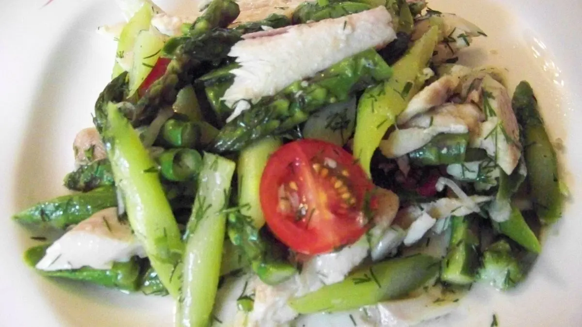 Ein frischer Salat aus gr&uuml;nem Spargel und ger&auml;ucherter Forelle liegt auf einem Teller, garniert mit Tomatenscheiben.