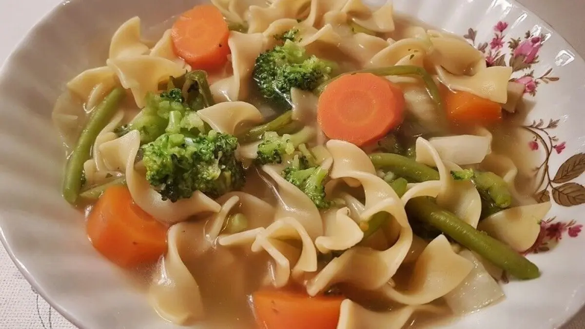 Eine Sch&uuml;ssel H&uuml;hnersuppe mit Bandnudeln, Karotten und Brokkoli, serviert auf einem gedeckten Tisch.