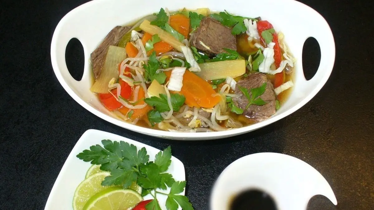 Eine Sch&uuml;ssel asiatische Rindfleisch-Gem&uuml;sesuppe steht auf einem Tisch, umgeben von frischen Kr&auml;utern und Limetten.