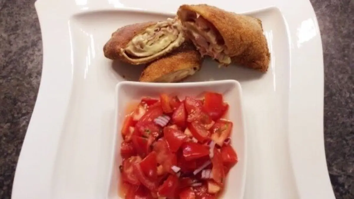 K&ouml;stliche Palatschinken-Cordon-Bleu auf einem Teller, neben einer Schale mit frischen, gehackten Tomaten.