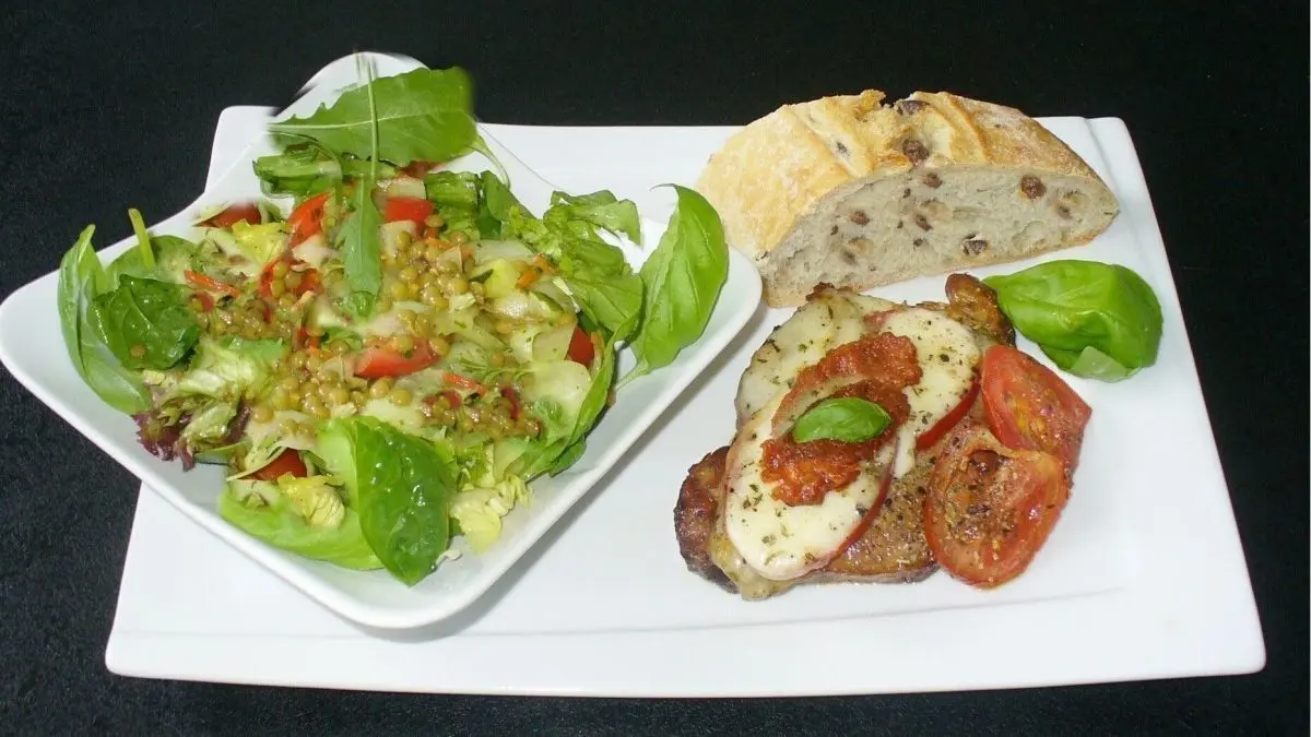 &Uuml;berbackenes Schweiner&uuml;ckensteak mit Mozzarella und Tomaten liegt auf einem wei&szlig;en Teller, begleitet von frischem Salat und Brot.