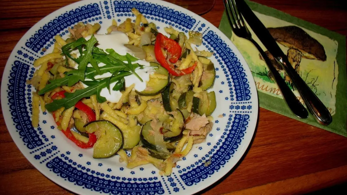 Eine bunte Gem&uuml;se-Sp&auml;tzle-Pfanne mit Thunfisch auf einem Teller, garniert mit Kr&auml;utern und serviert auf einem Tisch.