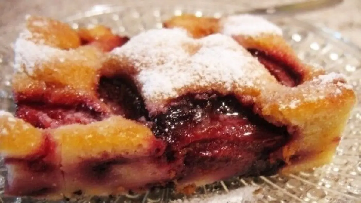 Ein St&uuml;ck Obstkuchen vom Blech liegt auf einem gl&auml;sernen Teller, best&auml;ubt mit Puderzucker und umgeben von K&uuml;chenutensilien.