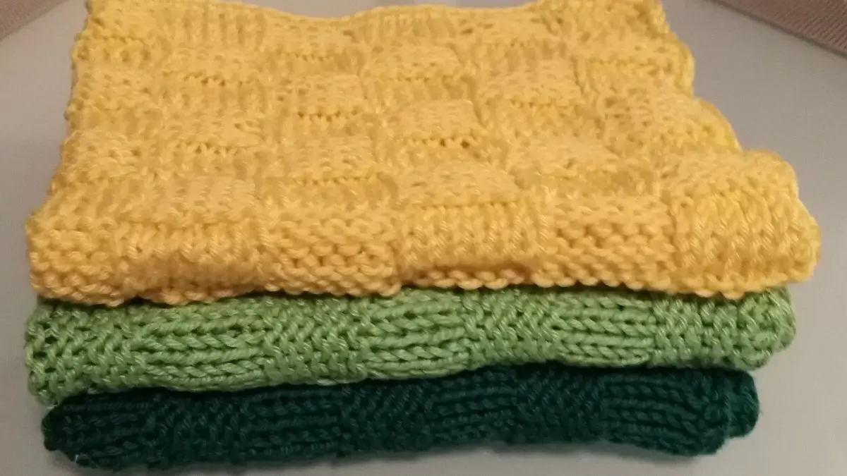 Gestapelte, handgestrickte Sp&uuml;lt&uuml;cher in Gelb, Gr&uuml;n und Dunkelgr&uuml;n liegen auf einer glatten Oberfl&auml;che.