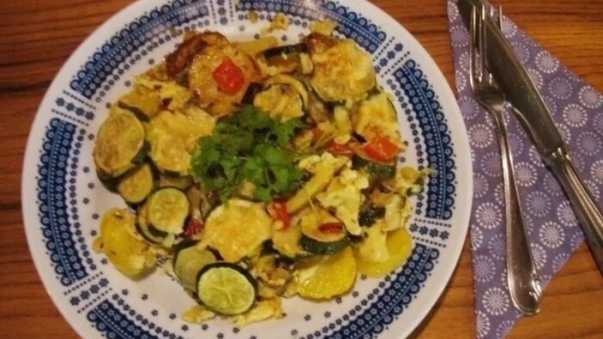 Ein bunter Zucchini-Paprika-Kartoffel-Auflauf mit goldbrauner K&auml;sekruste liegt auf einem Teller, umgeben von Besteck.