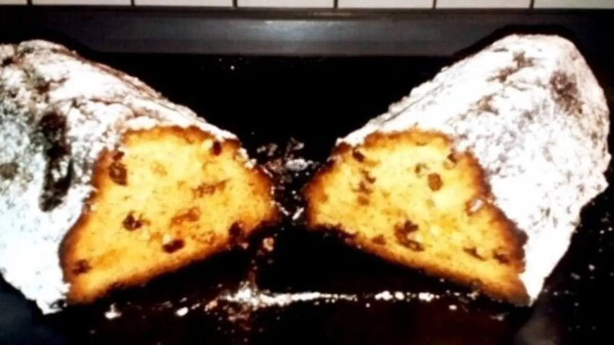 Ein Stollen mit einer goldenen, geschnittenen F&uuml;llung aus Quark und Marzipan liegt auf einem Teller, umgeben von Puderzucker.