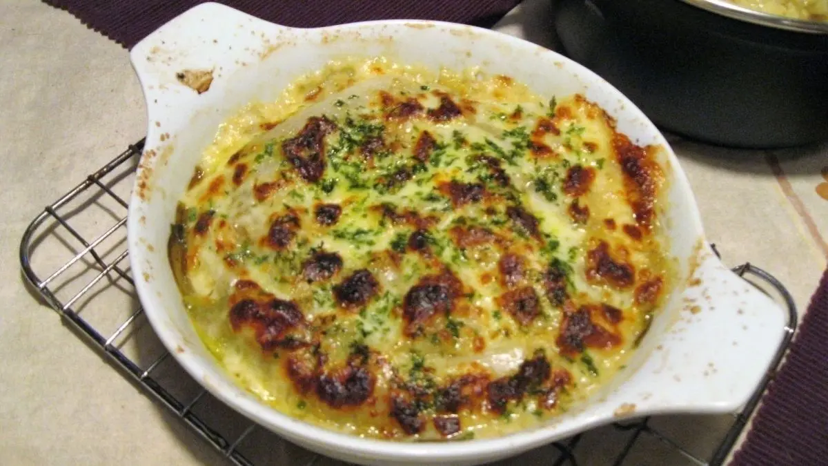 Ein goldbraun &uuml;berbackenes Chicoree-Gratin wird in einer wei&szlig;en Auflaufform pr&auml;sentiert, umgeben von einem rustikalen Tischsetting.