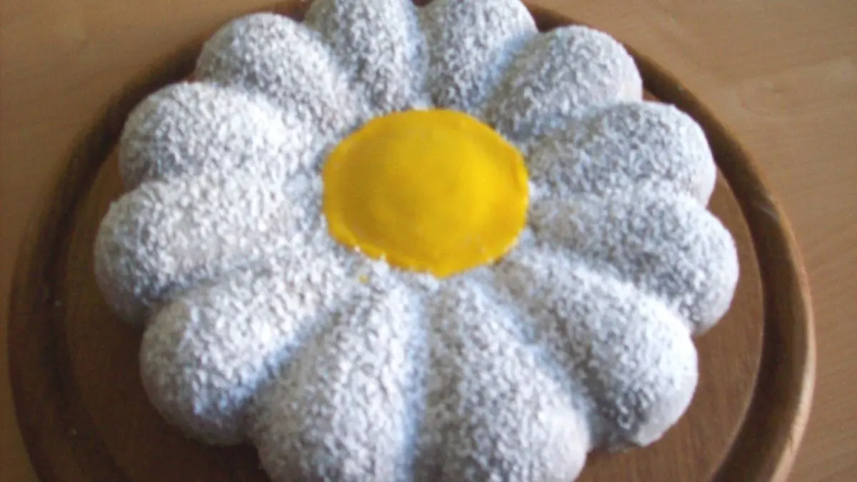 Eine blumenf&ouml;rmige Backform zeigt einen bunten Blumenkuchen mit gelbem Zentrum und wei&szlig;en Bl&uuml;tenbl&auml;ttern.
