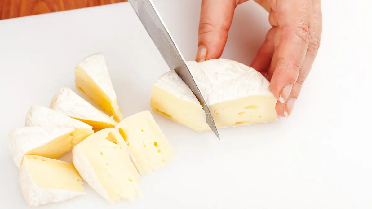 Ein handgeschriebener K&auml;se wird mit einem Messer auf einem Schneidebrett in gleichm&auml;&szlig;ige St&uuml;cke geschnitten.