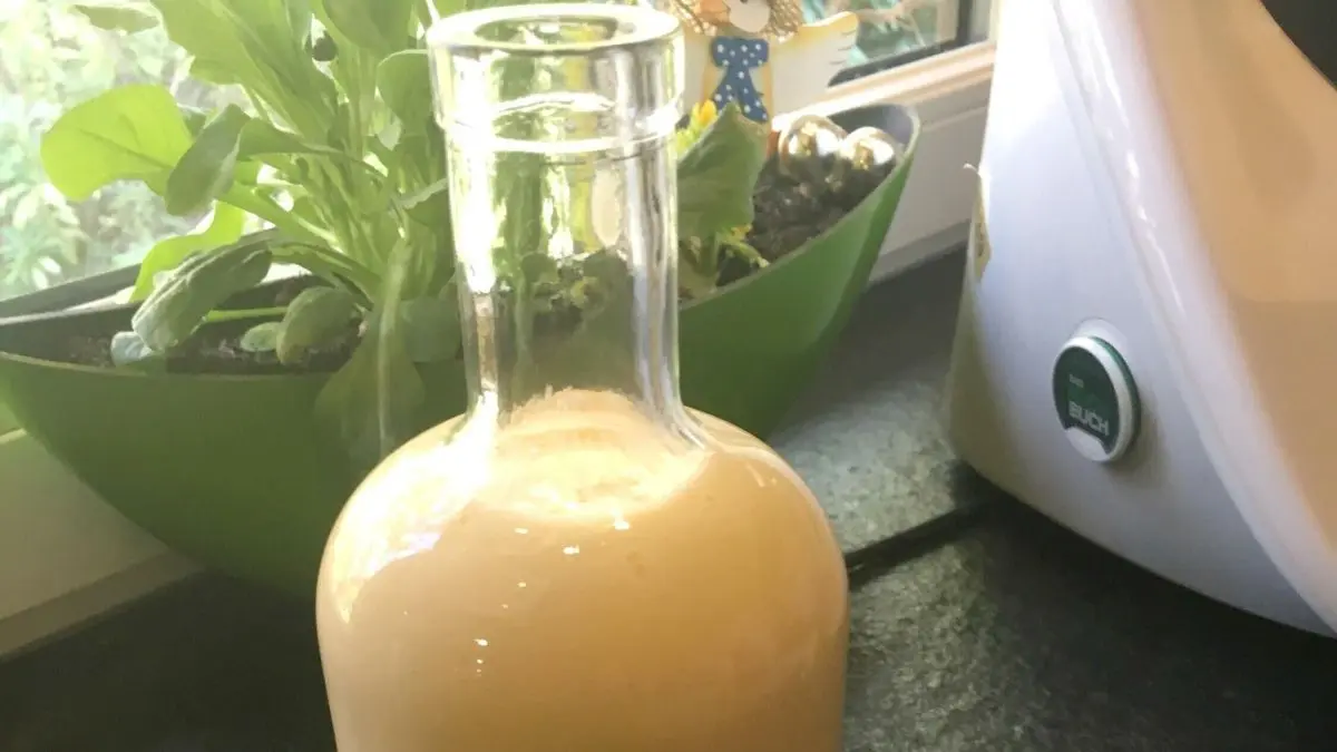 Eine Glasflasche mit cremigem Eierlik&ouml;r steht auf einer dunklen Oberfl&auml;che, umgeben von frischen Pflanzen und einem Thermomix.